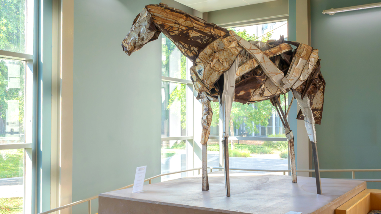 A sculpture of a horse made out of metal standing tall on a platform inside a building, looking out the window.